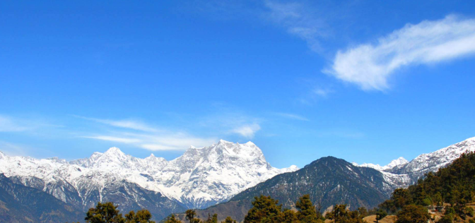 Scenic view of Chopta Valley with snow-capped Himalayan peaks and dense pine forests under a clear blue sky.